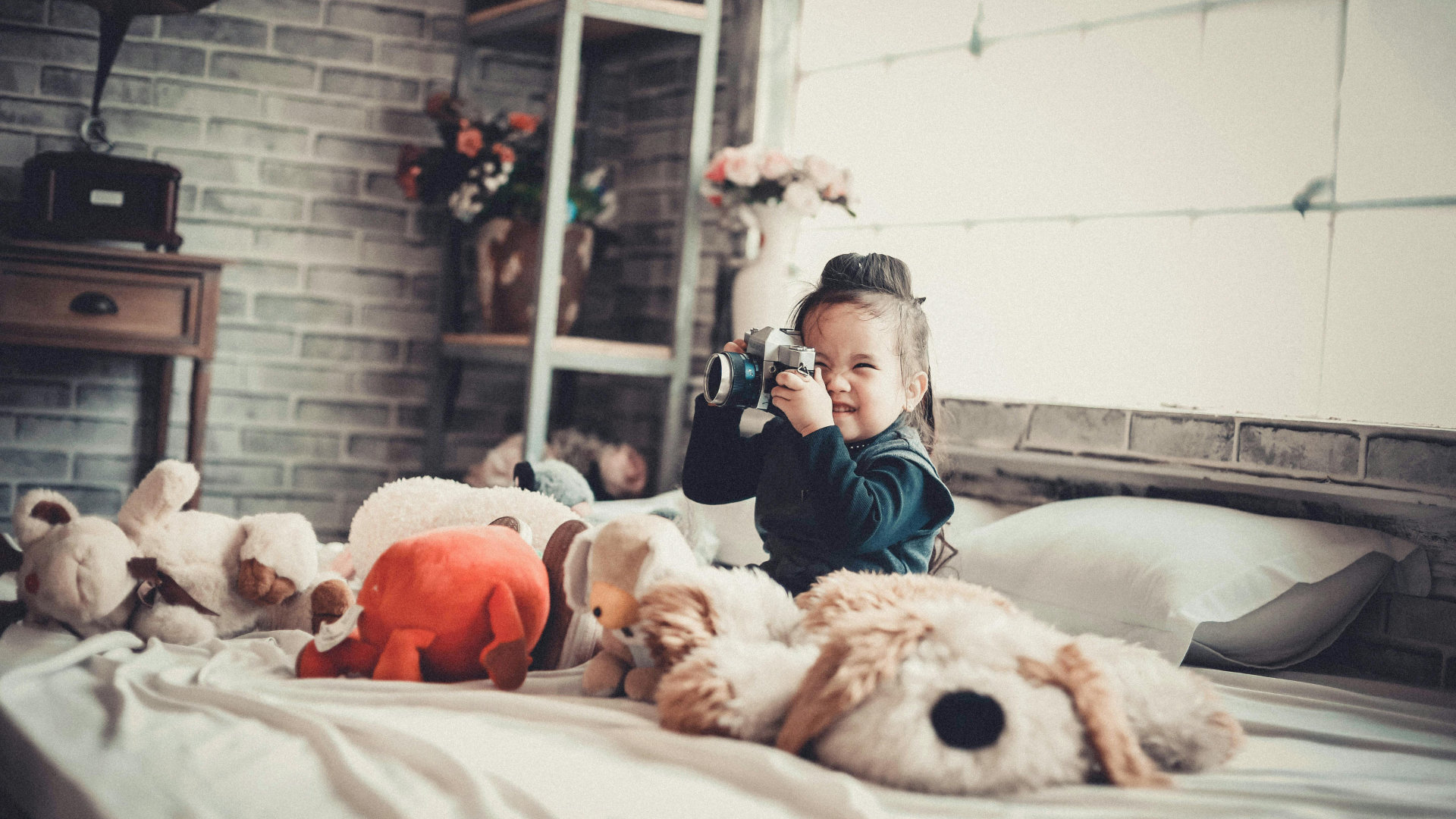 Child sits on bed playing with a camera surrounded by stuffed animals